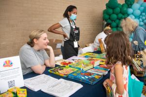 Three staff members in T shirts engage with customers across a table filled with colorful picture books.